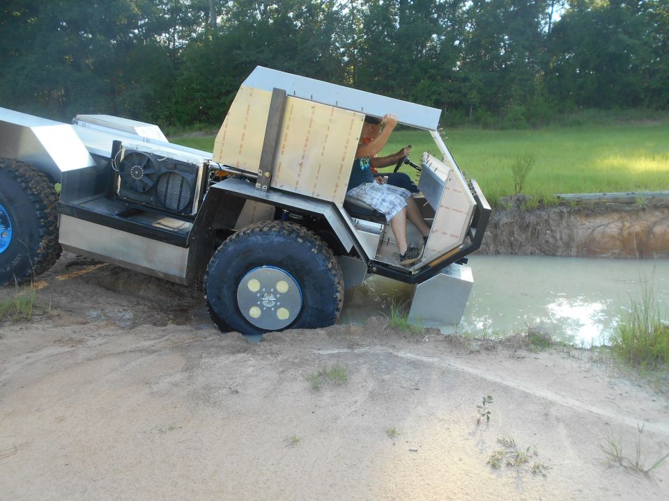 Custom amphibious vehicle with large tires partially in water, driver visible.