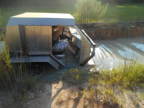 1992 Ford Bronco - A custom-built, boxy vehicle with a metallic exterior is partially submerged in muddy water, with a person inside operating the steering wh…