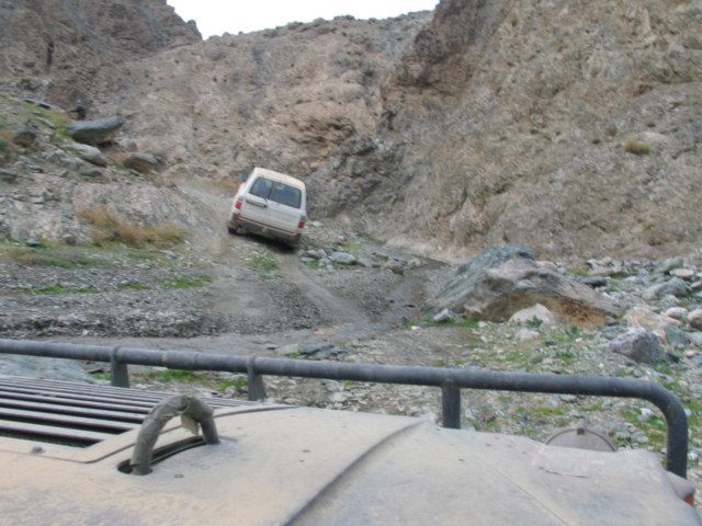 White van on a steep, rocky mountain road. Foreground shows part of another vehicle