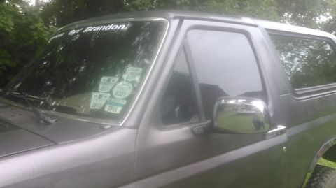 1992 Ford Bronco - Close-up view of the driver's side of a dark gray Ford Bronco. The windshield has several stickers, including one that reads "By Brandon".
