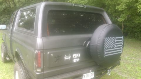1992 Ford Bronco - The rear passenger side of a dark gray SUV is shown parked on a grassy area with trees in the background. A spare tire cover with a wrench-…