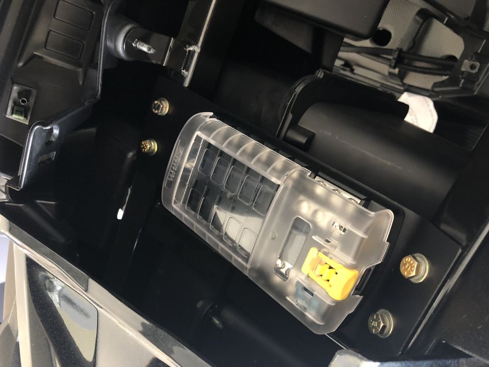 Close-up of a clear plastic fuse box with a yellow lever inside, mounted in a black vehicle interior.