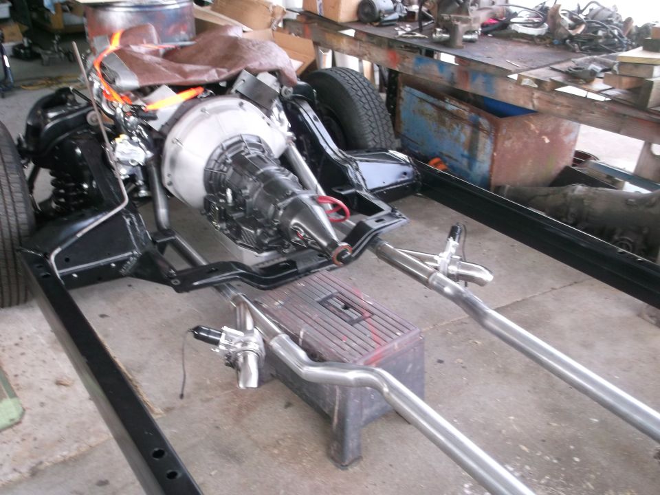 Car chassis with engine and transmission installed, showing exhaust pipes and suspension components
