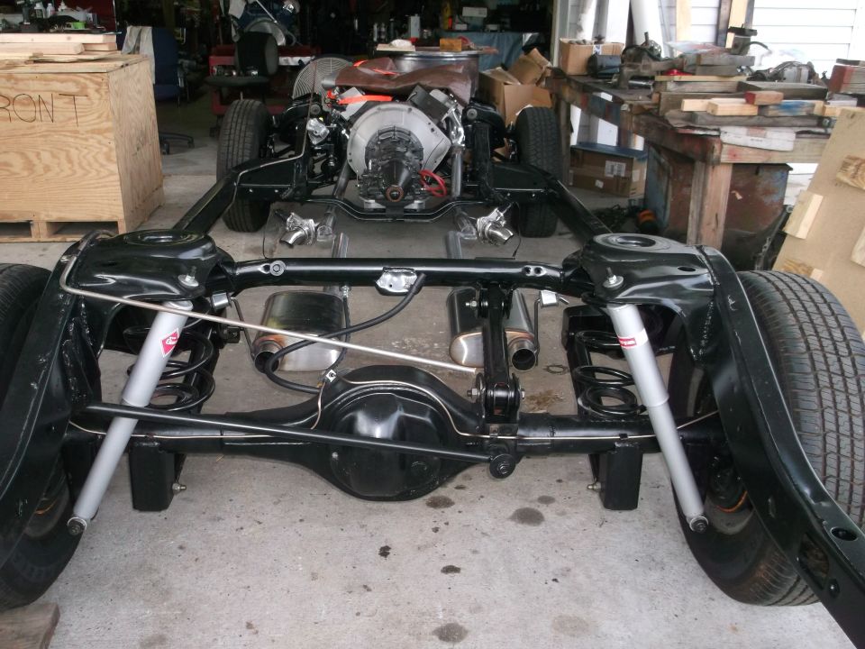 Car chassis with engine, rear axle, suspension, and tires visible. Some tools and wooden crates in background