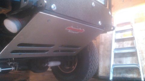 1992 Ford Bronco - Low-angle underside view of a bolt-on front skid plate mounted beneath a lifted truck, finished in gray with louvered cooling slots and a r…