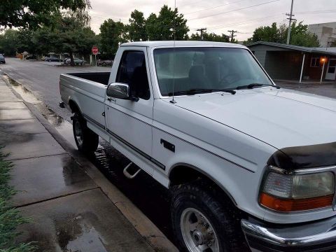 1995 Ford Bronco - This white pickup truck is parked on a wet street next to a sidewalk, with trees and houses in the background.