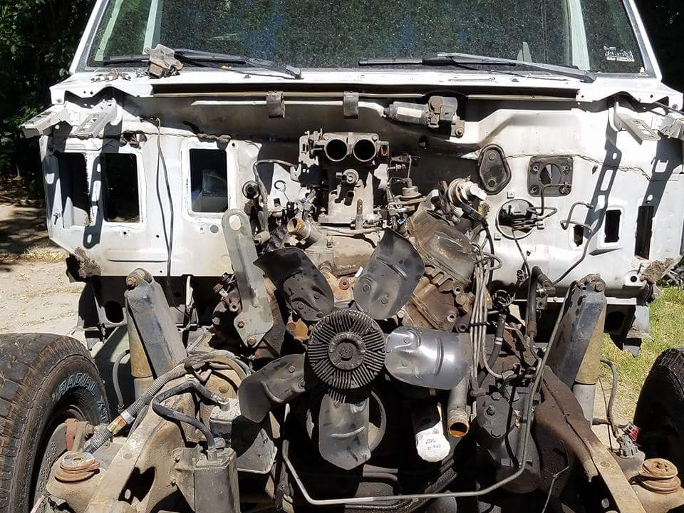 Front end of a vehicle with engine exposed, showing the fan, carburetor, and chassis components. The bodywork is damaged and removed