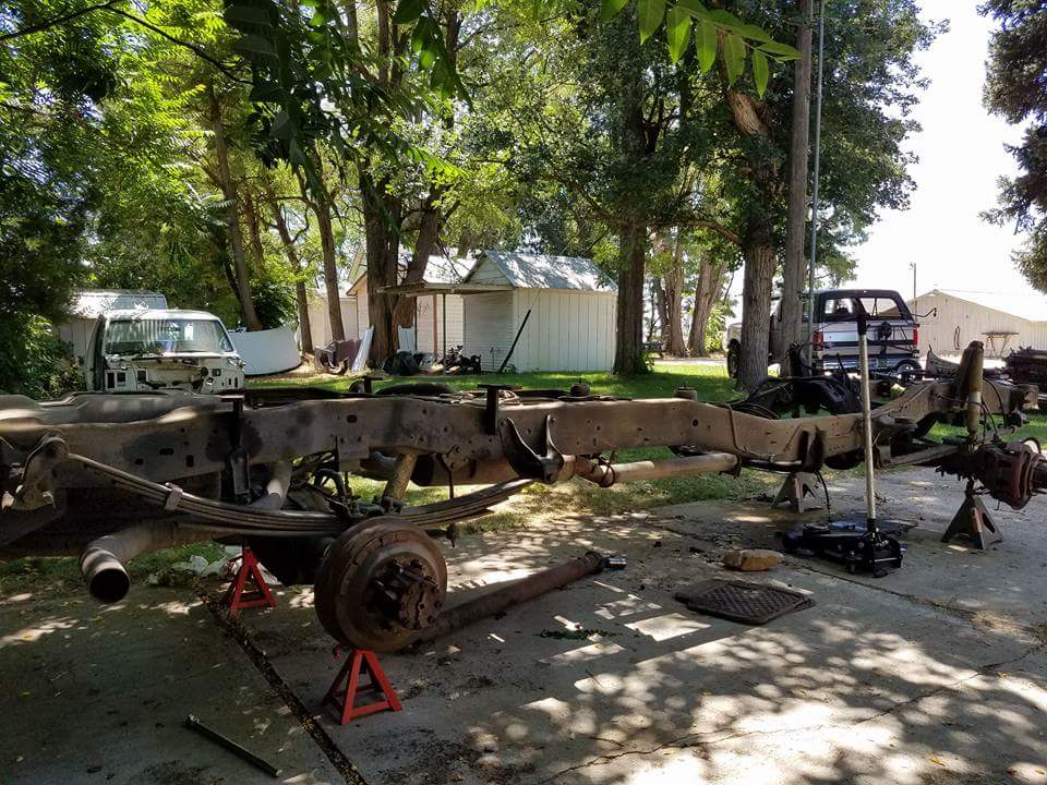 Truck frame disassembled and supported by jack stands outdoors, with a partially visible truck cab in the background