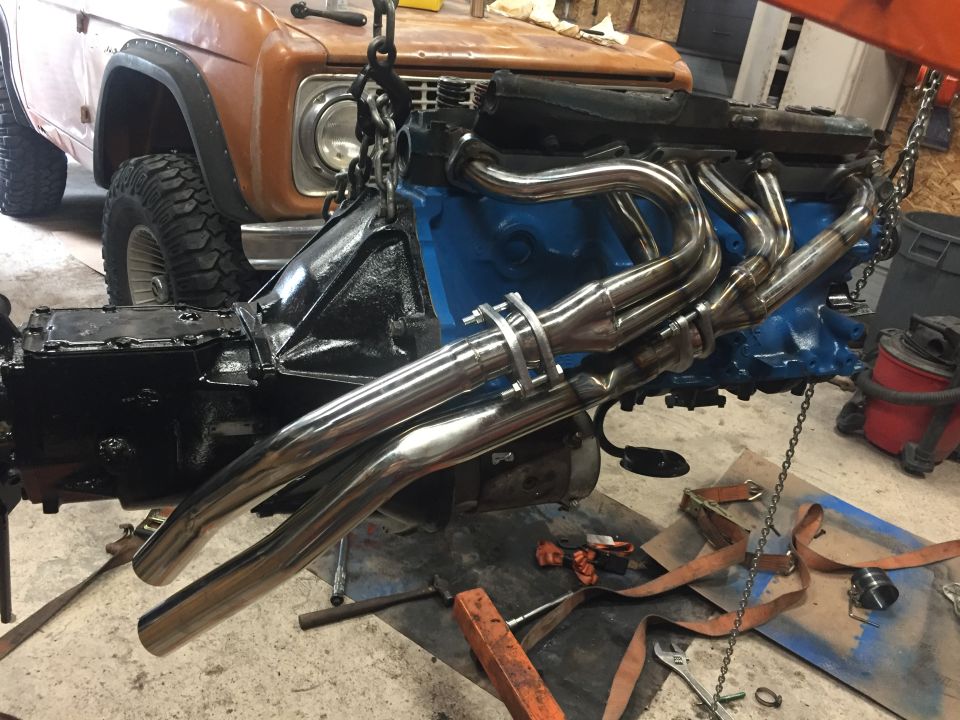 Engine and transmission assembly with chrome headers being lowered by chains. Partially visible 1969 Ford Bronco front end in background