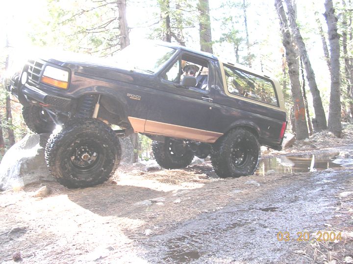 Lifted black and tan Ford Bronco flexing over a boulder on a forest trail