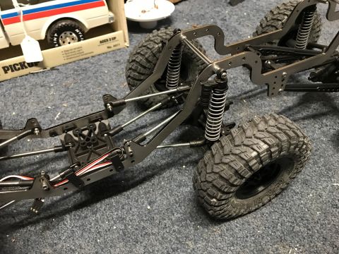 1989 Ford Bronco - Close-up of an RC crawler chassis with large tires and suspension components. A toy pickup truck box is visible in the background.