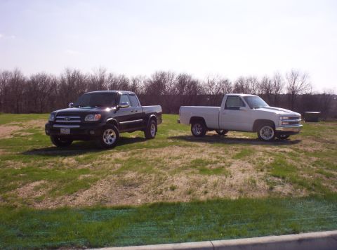 1988 Ford Bronco - Two pickup trucks, a black Toyota Tundra and a white Chevrolet, are parked side-by-side on a grassy field under a clear sky.