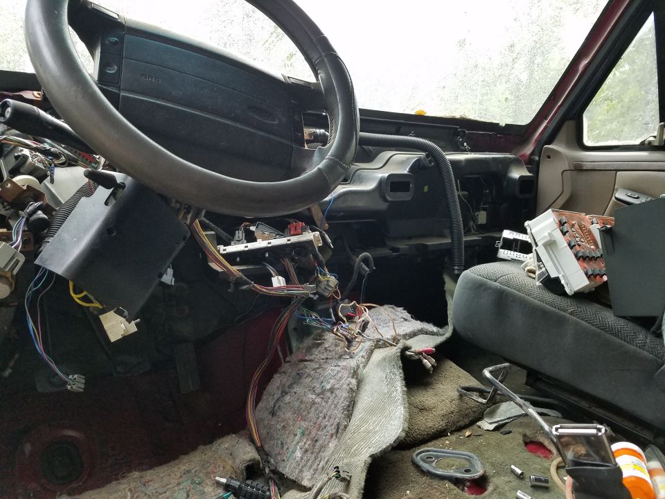 Torn-apart truck interior with dash removed, exposed wiring harnesses, steering wheel intact, insulation on floor