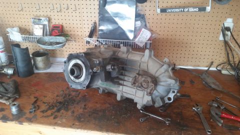1985 Ford Bronco - A disassembled transfer case sits on a cluttered workbench, surrounded by tools and automotive parts.