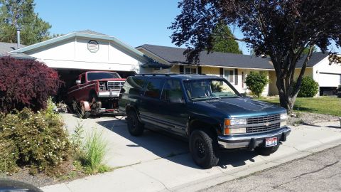 1981 Ford Bronco - A mid-1990s Chevrolet Suburban is parked on a driveway, with a red pickup truck on a trailer being towed into the garage behind it.
