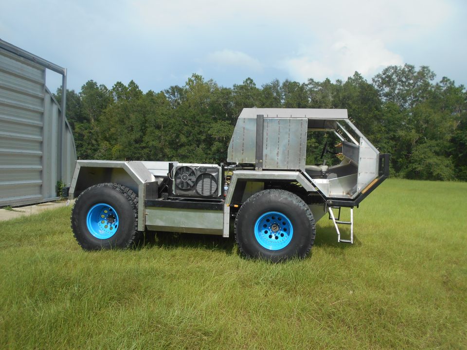 Custom all-terrain vehicle with blue wheels, exposed engine, and aluminum body, parked on grass
