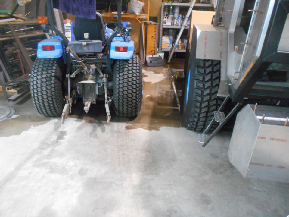 Rear view of a blue tractor next to a metal trailer. Tractor has large rear tires and a tow hitch