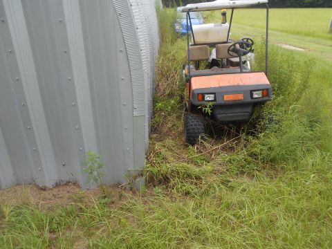 1992 Ford Bronco - An orange golf cart sits abandoned in overgrown grass next to a corrugated metal structure.