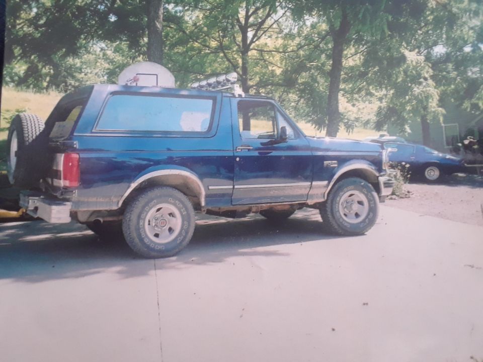 Blue 1994 Ford Bronco parked on concrete driveway with spare tire on rear door. Another car is visible in the background