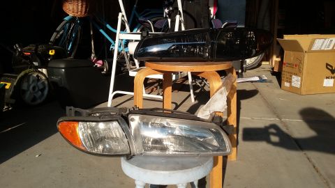 2000 Pontiac Grand Prix - Pair of clear-lens headlight assemblies on the floor and a black rear spoiler resting on a stool in a cluttered garage.