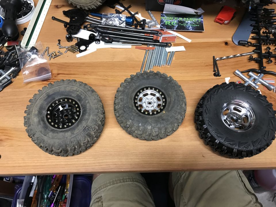 Three RC car tires and wheels on a wooden surface with tools and parts.