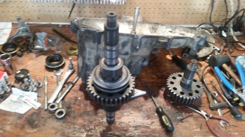 1985 Ford Bronco - A workbench is covered in transmission parts and tools. A large metal casing is in the background, with gears and shafts laid out in the fo…