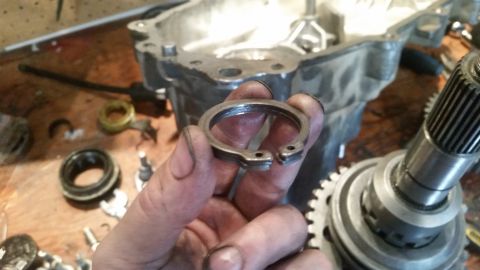 1985 Ford Bronco - A hand holds a metal snap ring with two holes for pliers, in front of a disassembled transmission housing and a gear shaft.