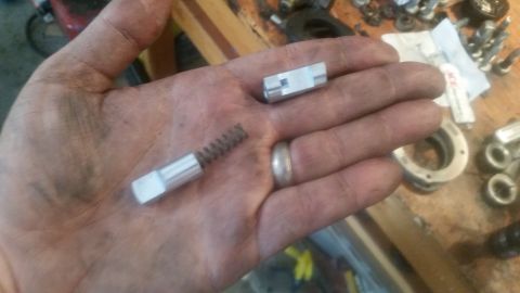 1985 Ford Bronco - A person's dirty hands hold two small metal parts, one with a spring attached. The background shows a workbench with various tools and part…