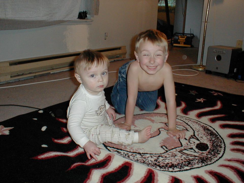 Two young boys on a rug with a moon and sun design. The older boy is shirtless and on his knees, smiling. The younger boy is wearing a long-sleeved shirt and pants