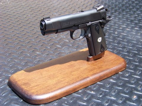 1995 Mercury Grand Marquis - A black semi-automatic pistol with "COLT GUNSITE PISTOL" engraved on the slide rests on a wooden display stand. The pistol is angled upward…