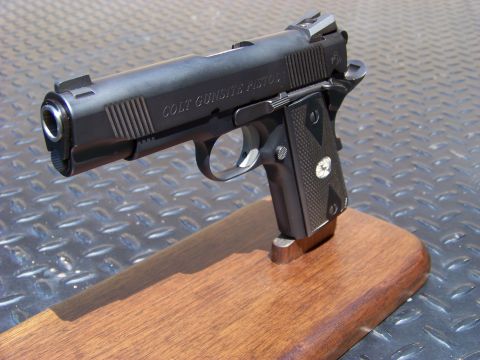 1995 Mercury Grand Marquis - A black Colt Gunsite Pistol is displayed on a wooden stand. The pistol is angled to the left, showing the slide with "COLT GUNSITE PISTOL"…