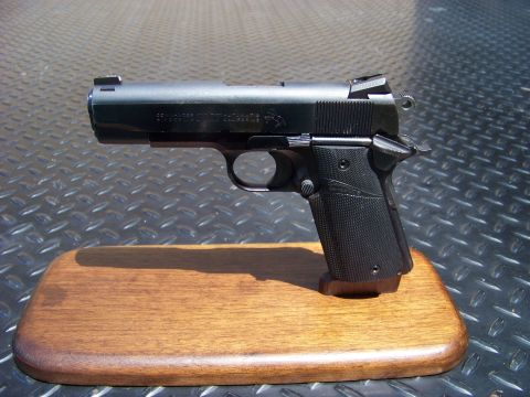 1995 Mercury Grand Marquis - A black semi-automatic pistol is displayed on a wooden stand. The pistol has "COMMANDER .45 AUTO" and "COLT" markings visible on the slide.