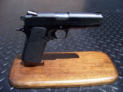 1995 Mercury Grand Marquis - A close-up, angled shot of a black semi-automatic pistol resting on a wooden display stand. The pistol is positioned on a dark, textured su…