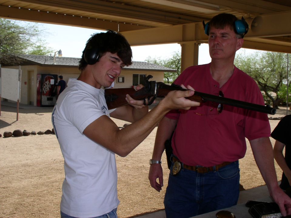 Young man aims rifle with instructor watching at outdoor shooting range