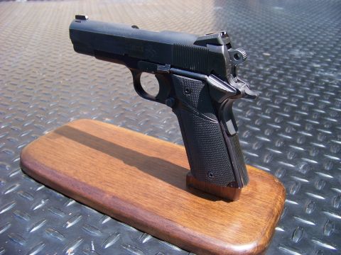 1995 Mercury Grand Marquis - A black semi-automatic pistol is displayed on a wooden base. The pistol is angled upwards and to the left, with its shadow cast on the base.
