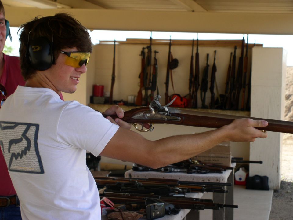 Man wearing ear protection and yellow safety glasses aims an antique rifle. Several other rifles are displayed on a shelf behind him