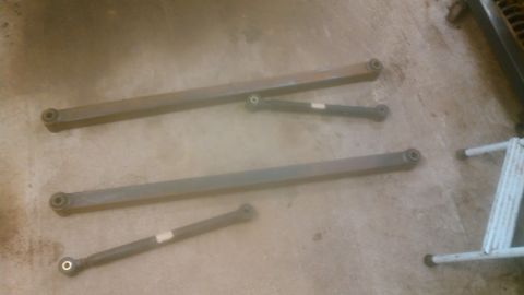 1992 Ford Bronco - Two sets of metal control arms are laid out on a concrete floor. Each set consists of a longer arm and a shorter arm, both with eyelet ends.