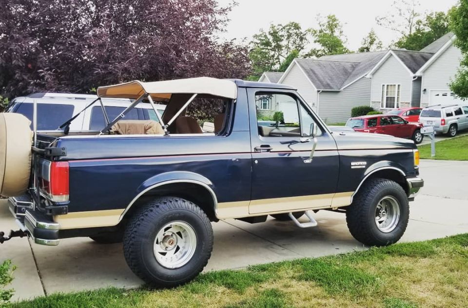 1989 Ford Bronco with tan soft top and spare tire on rear, parked on driveway.