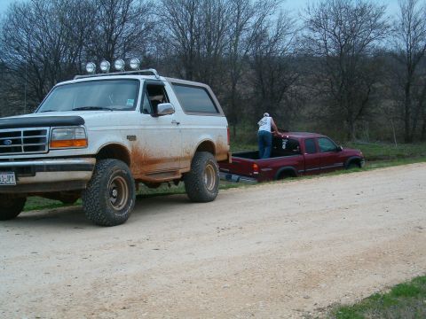 1996 Ford Bronco - A white 1996 Ford Bronco tows a maroon pickup truck that is stuck in a ditch on a dirt road.
