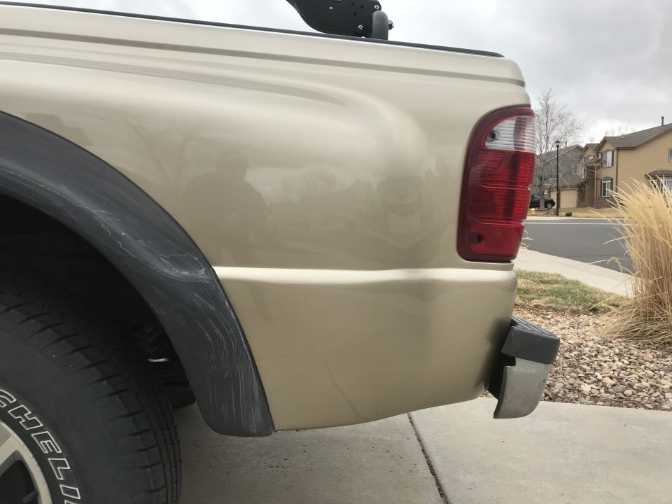 Rear passenger side corner of a gold pickup truck, showing the taillight, bumper corner, and fender flare