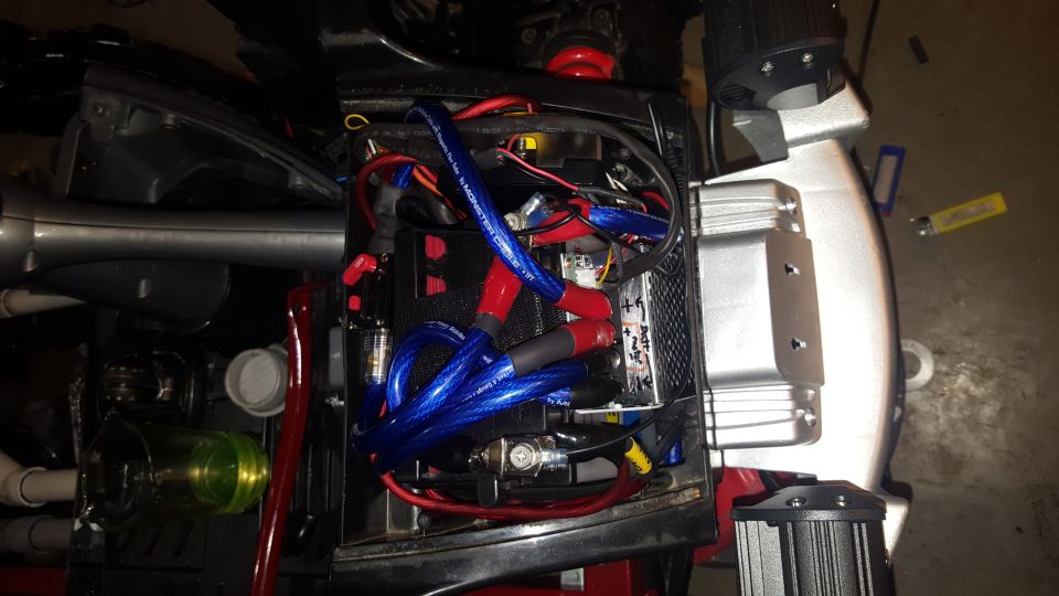 Vehicle battery compartment with blue and red wires, a battery, and a silver component. Some text is visible on the battery