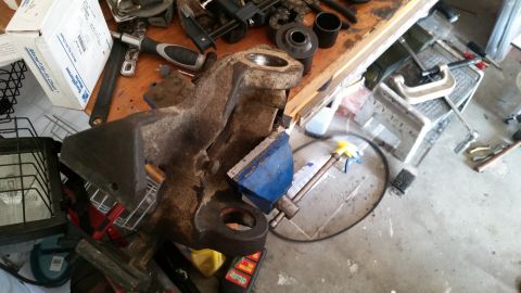 1985 Ford Bronco - A close-up view of a rusty metal automotive part held in a blue vise on a workbench. Various tools and a box are scattered around the workb…