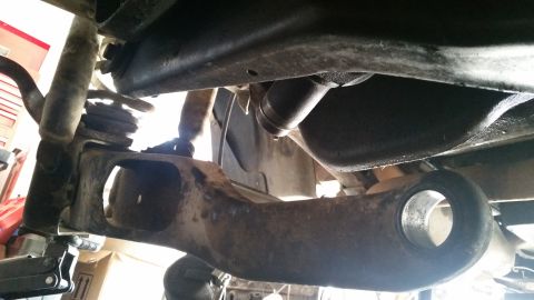 1985 Ford Bronco - Underneath view of the suspension components of a 1985 Ford Bronco, showing a rusty control arm and shock absorber assembly. The vehicle ap…