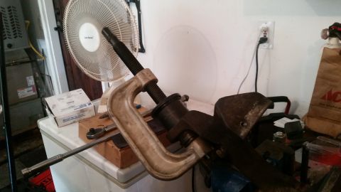 1985 Ford Bronco - A close-up shot of a C-clamp tool pressing on a black cylindrical part, set against a white wall with an electrical outlet. Tools and boxes…