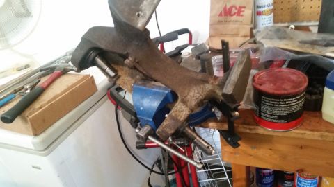 1985 Ford Bronco - A rusty vehicle part is clamped in a blue vise on a workbench. Various tools and cans of lubricant are visible in the background.