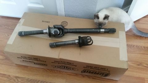1985 Ford Bronco - Two driveshafts with U-joints and splined ends are placed on top of a cardboard box. A Siamese cat is in the background, looking at the dri…