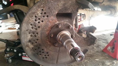 1985 Ford Bronco - A close-up view of a heavily rusted and dirty brake rotor and hub assembly, likely from a 1985 Ford Bronco, supported by a black hydraulic…