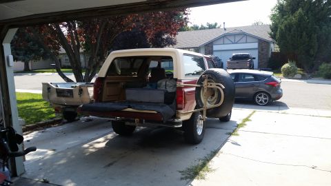 1985 Ford Bronco - The rear of a two-tone red and tan Ford Bronco is shown parked under a carport. The tailgate is down, revealing cargo inside, and a spare t…