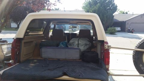 1985 Ford Bronco - The rear cargo area of a tan and red SUV is filled with various items, including bags, a cooler, and a blanket. A spare tire is mounted on…
