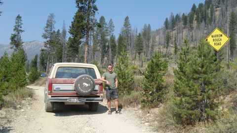1985 Ford Bronco - A man stands next to a red and tan Ford Bronco on a dirt road with a "Narrow Rough Road" sign in the background. The Bronco has a spare tir…